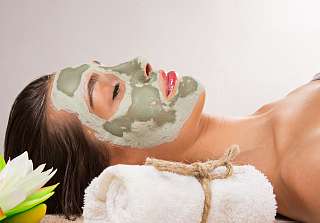 Femme détendue portant un masque à l’argile verte, allongée sur une serviette blanche roulée, à côté d’une fleur de lotus