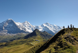 Groupe de randonneurs debout sur une crête herbeuse du Männlichen avec une vue panoramique spectaculaire sur l'Eiger, le Mönch et la Jungfrau sous un ciel bleu.