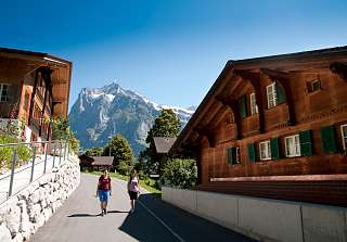 Two walkers on a village street in Grindelwald, surrounded by traditional Swiss wooden houses with views of the snow-capped Wetterhorn – Sunstar Hotel Grindelwald.