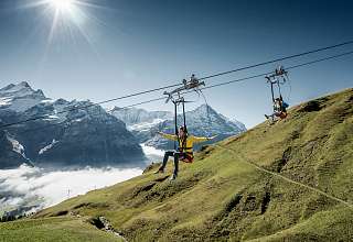 Deux personnes sur une tyrolienne au-dessus de versants verdoyants avec vue sur la vallée de Grindelwald couverte de nuages et les sommets alpins environnants.