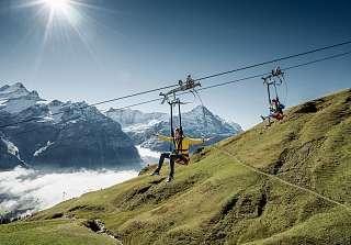 Two people on the First Flyer zipline above Grindelwald with a spectacular view of the snow-capped peaks of the Bernese Oberland – pure adrenaline at Sunstar Hotel Grindelwald.