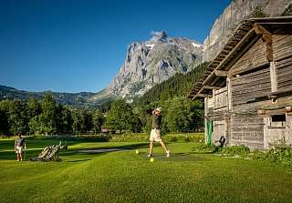 Golfer schlägt ab auf dem Golfplatz Grindelwald mit Blick auf das Wetterhorn und ein traditionelles Bergchalet