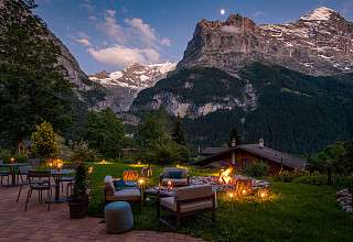 Terrasse extérieure chaleureusement éclairée avec fauteuils lounge, feu de camp et bougies devant un imposant panorama montagnard à Grindelwald au crépuscule et au clair de lune.