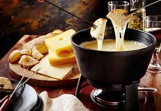 Fondue au fromage fumante dans un caquelon noir avec du fromage fondu sur une fourchette, servie avec des tranches de pain et du fromage sur une planche en bois.