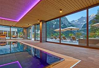 Innenpool im Sunstar Hotel Grindelwald mit violetter Ambientebeleuchtung, Hängesesseln und Panoramafenstern zur Terrasse und Bergkulisse