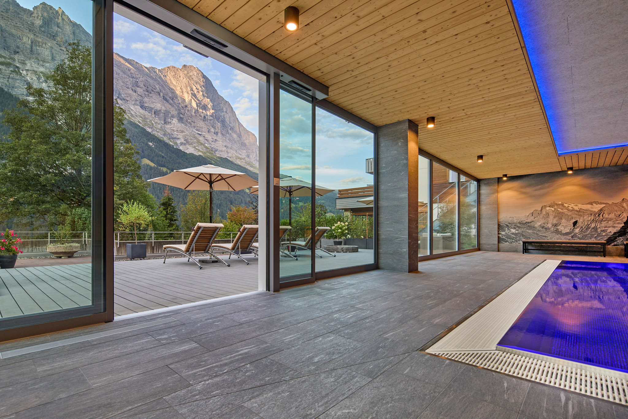 Open glass sliding door from the indoor pool of the Sunstar Hotel Grindelwald onto the sun terrace with loungers and parasols, with an imposing rock face in the evening light behind