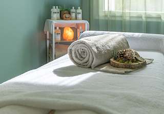 Bright treatment room with green walls, a prepared massage table with white towels and dried flower decoration, with a salt lamp and skincare products in the background