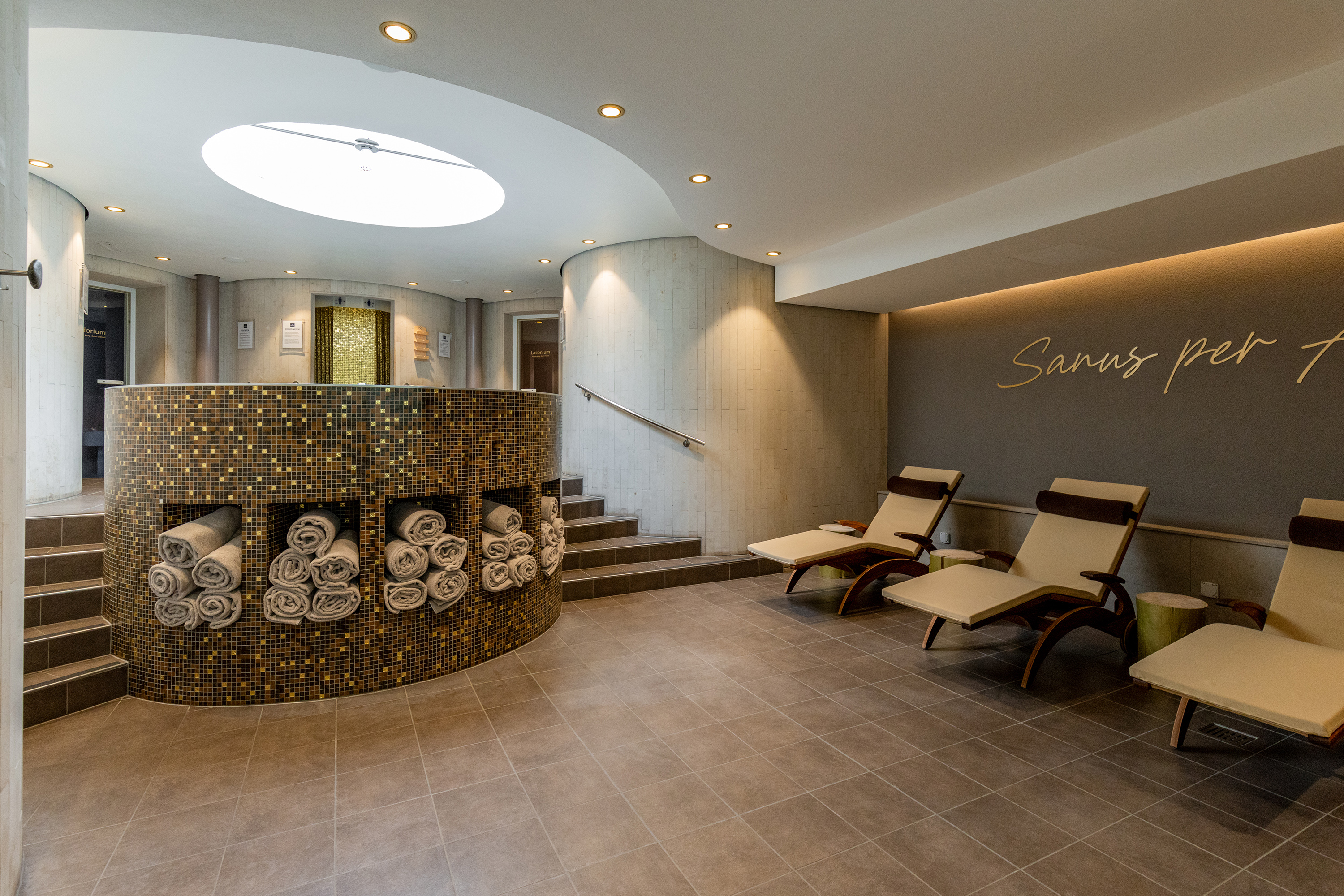 Zwei Personen in weissen Bademänteln gehen durch einen modernen Spa-Ruhebereich mit Mosaikwand, aufgerollten Handtüchern, Liegestühlen und der goldenen Schriftzug-Wand «Sanus per Aquam».