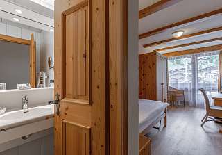 Vue depuis la salle de bains vers la chambre simple Classic avec porte en bois, lit simple, armoire et balcon avec vue sur le village de montagne